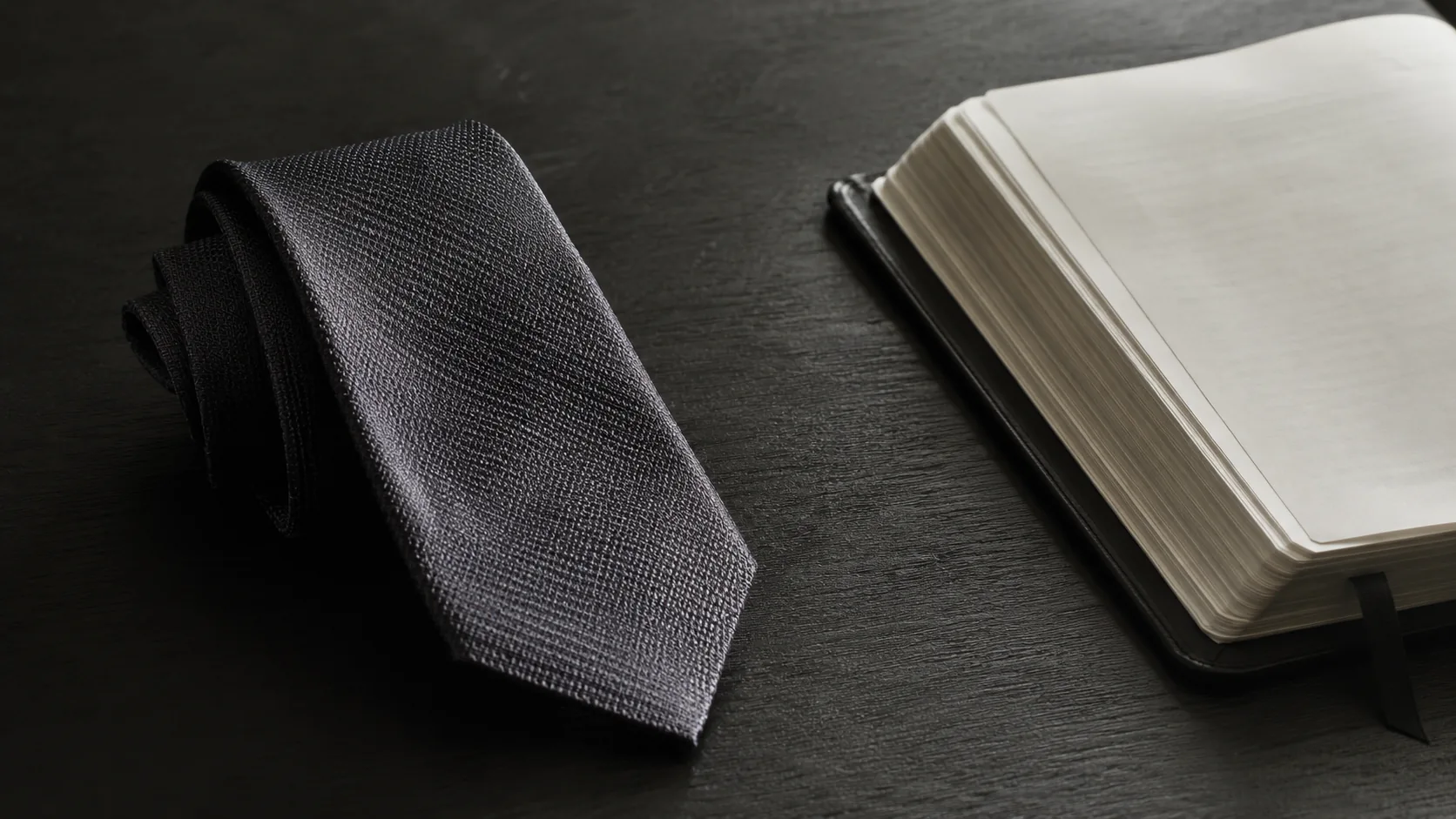 Corbata de seda doblada sobre una mesa oscura junto a un libro abierto, en una escena sobria sobre fragilidad humana y gracia.