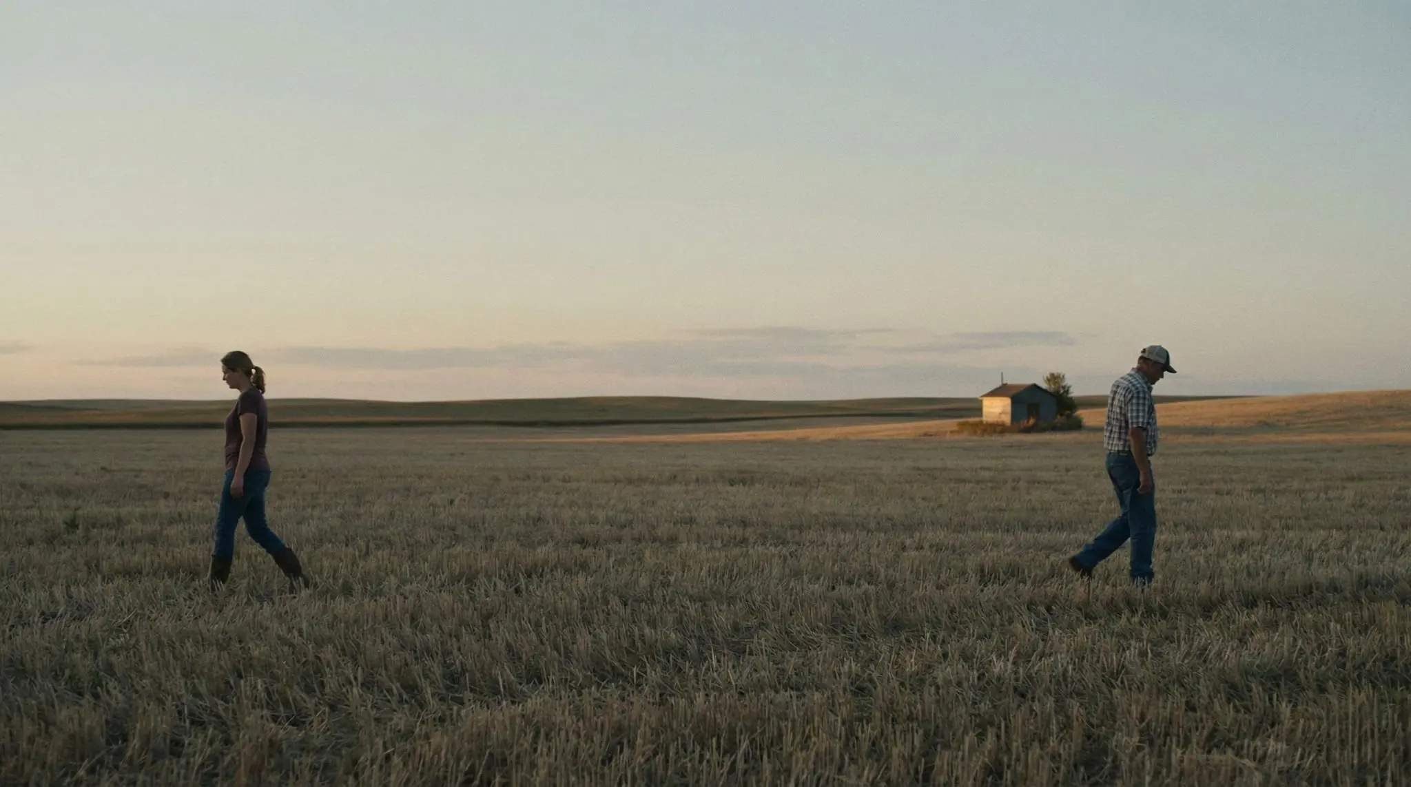 Dos personas caminan separadas en un campo de cosecha al atardecer, en una escena rural sobria que sugiere distancia, cuidado y refugio silencioso.