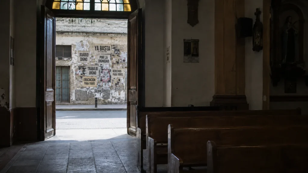 Interior de una iglesia con la puerta abierta hacia una calle cubierta de carteles sobre dignidad, derechos y justicia social.